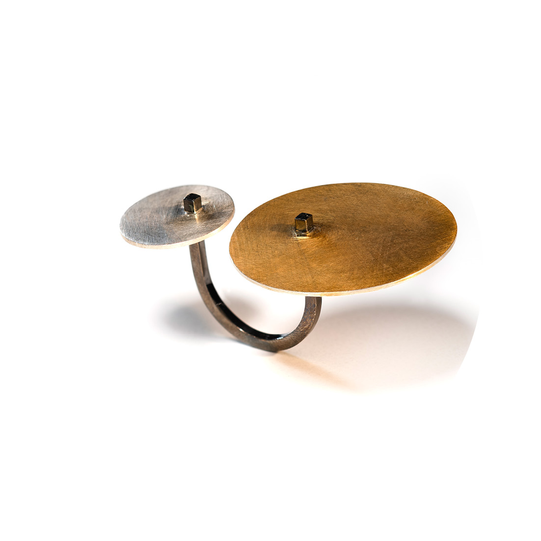 O anel Orbitas é feito com fio quadrado, banhado em ródio negro, e nele “orbitam” duas chapas circulares, uma grande com banho de ouro e outra pequena em prata fosca.