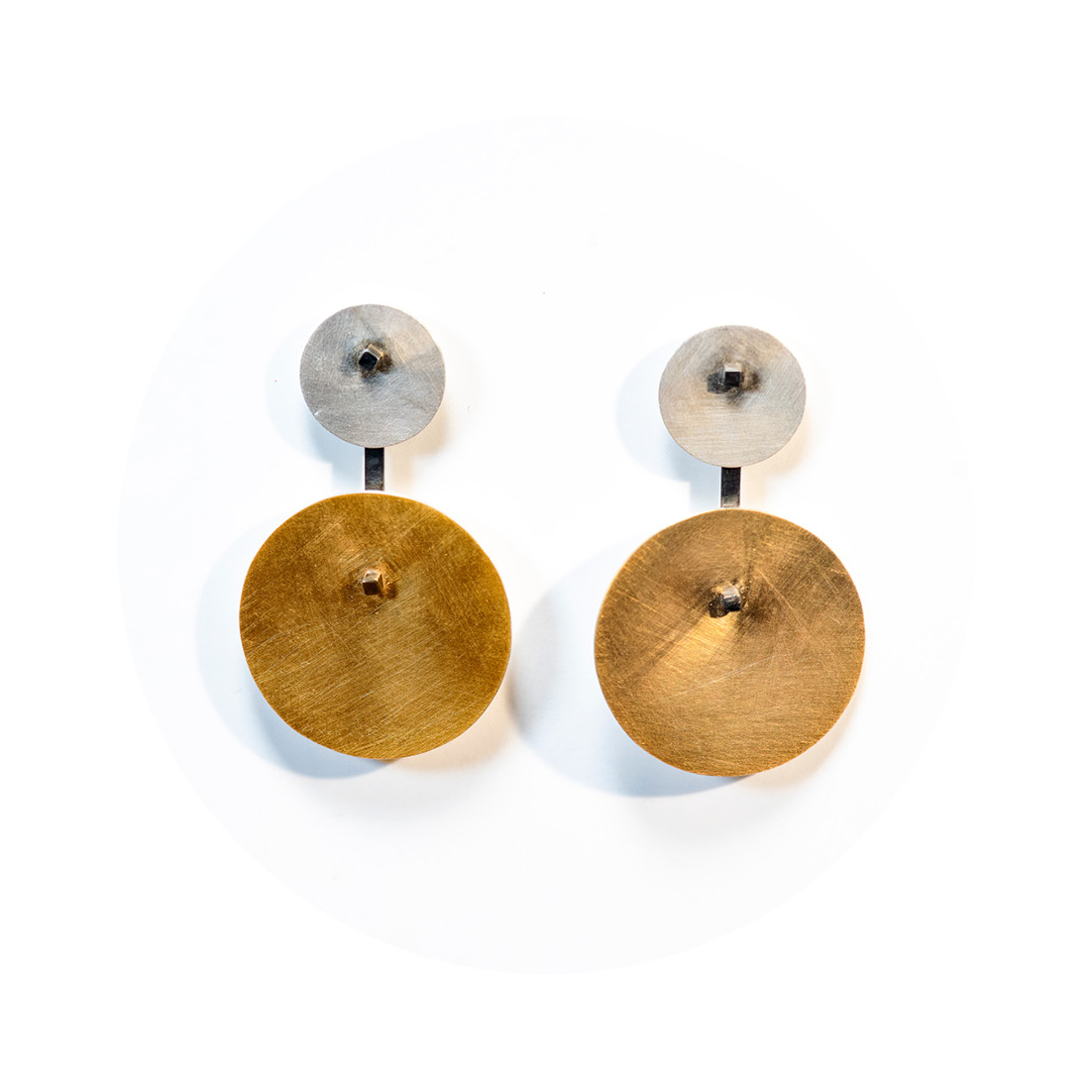 O Brinco una Orbitas tem duas chapas circulares, uma pequena em prata fosca, conectada a um pino quadrado e haste com banho de ródio, e outra grande com banho de ouro 750, que fica pendurada, conectada por esta haste.