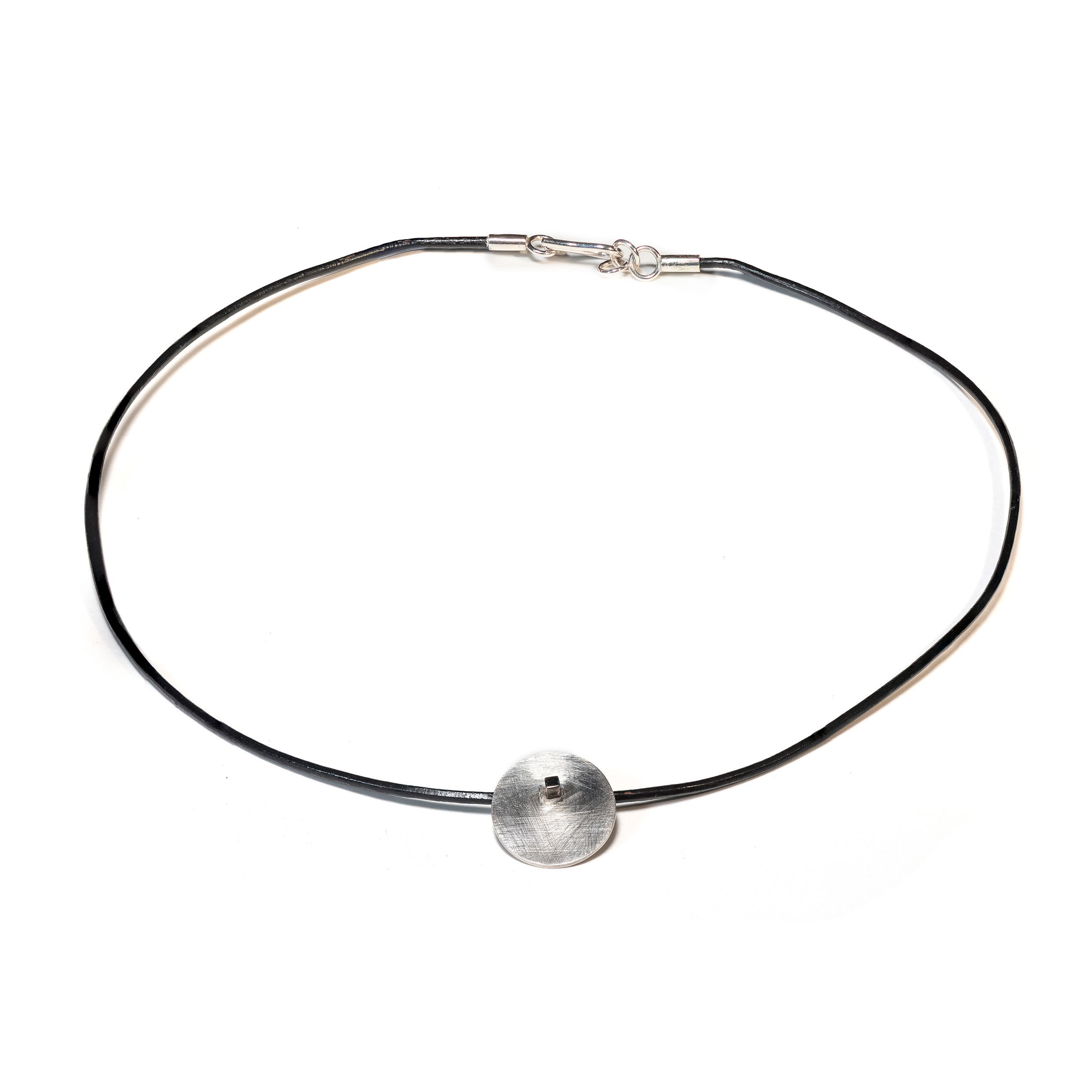 O Colar una Ar é feito com fio de couro preto, com pingente em chapa circular fosca, sustentada por um pino quadrado, banhado em ródio negro.