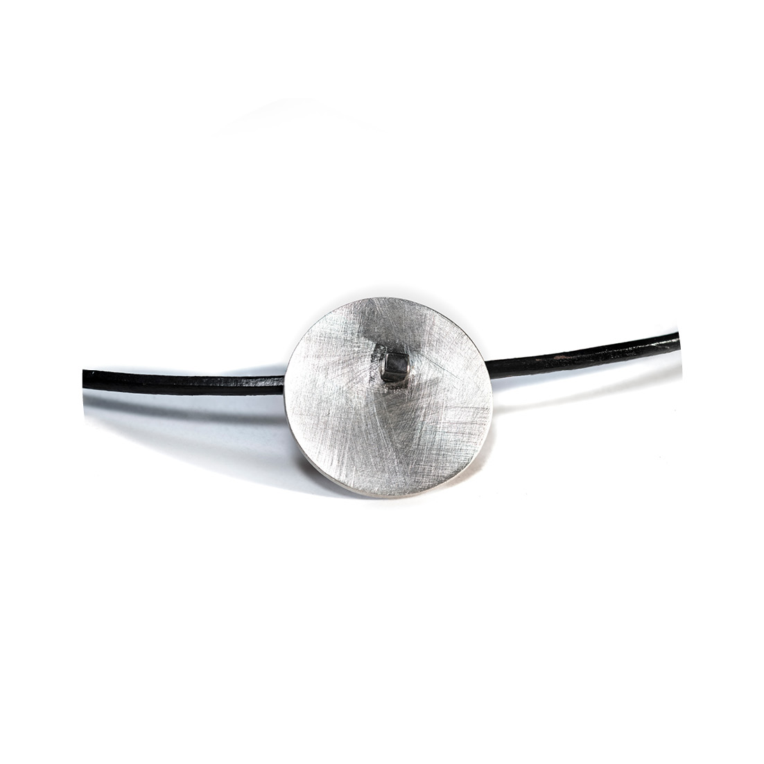 O Colar una Ar é feito com fio de couro preto, com pingente em chapa circular fosca, sustentada por um pino quadrado, banhado em ródio negro.